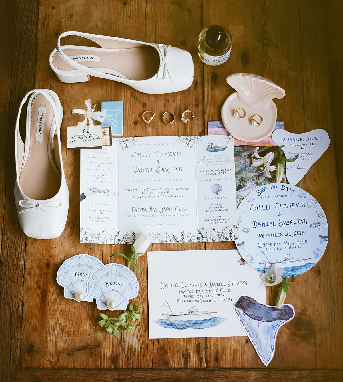A styled flat-lay of wedding stationery and accessories arranged on a wooden table, including white Mary Jane shoes, a shell dish with rings, invitation cards, napkins, and small floral accents.