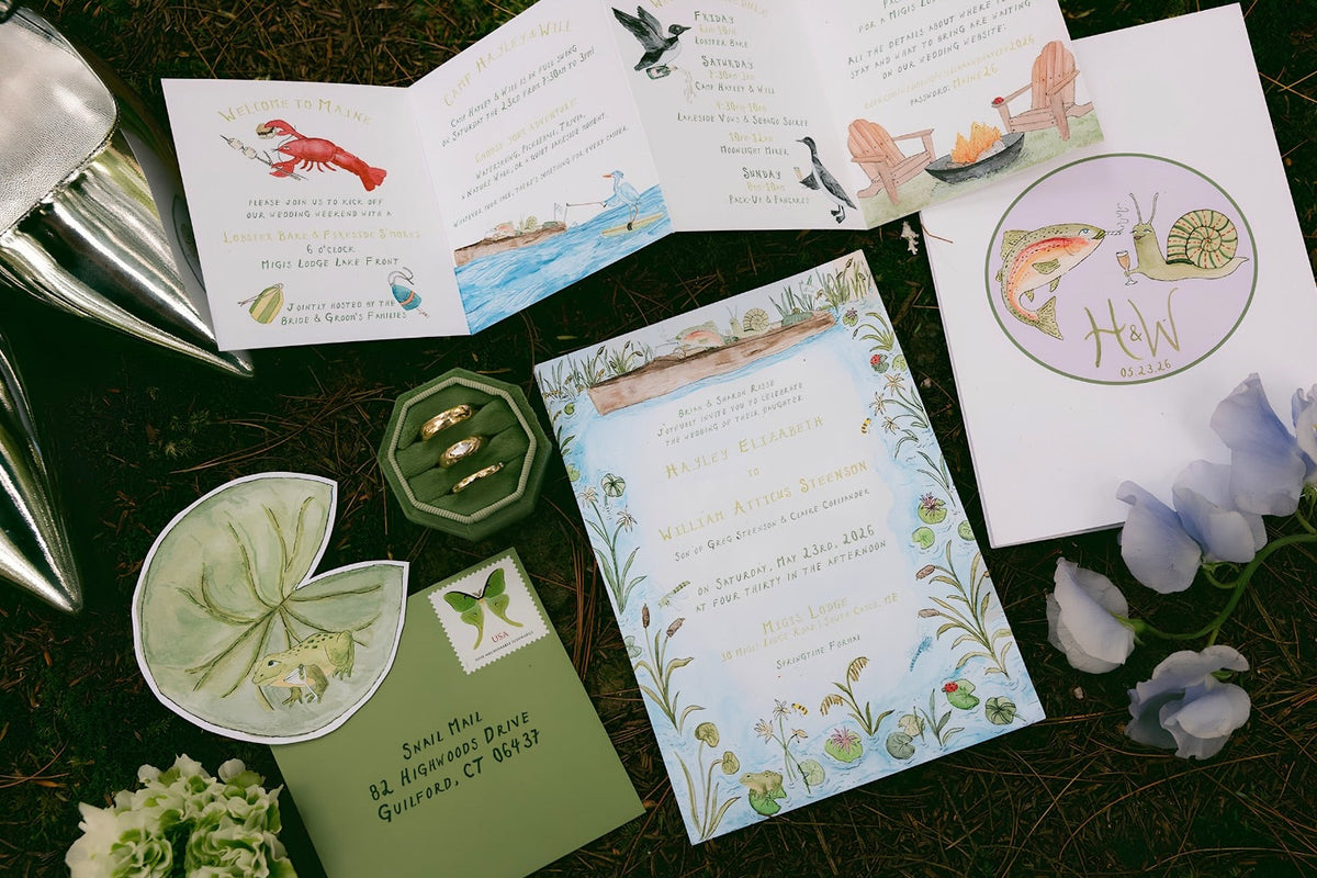 A styled flat-lay of a nature-themed wedding invitation suite on dark forest floor mulch, featuring a central floral invitation card, an open instruction booklet, a green envelope, a green octagonal box, a lily pad-shaped card, and decorative flowers.
