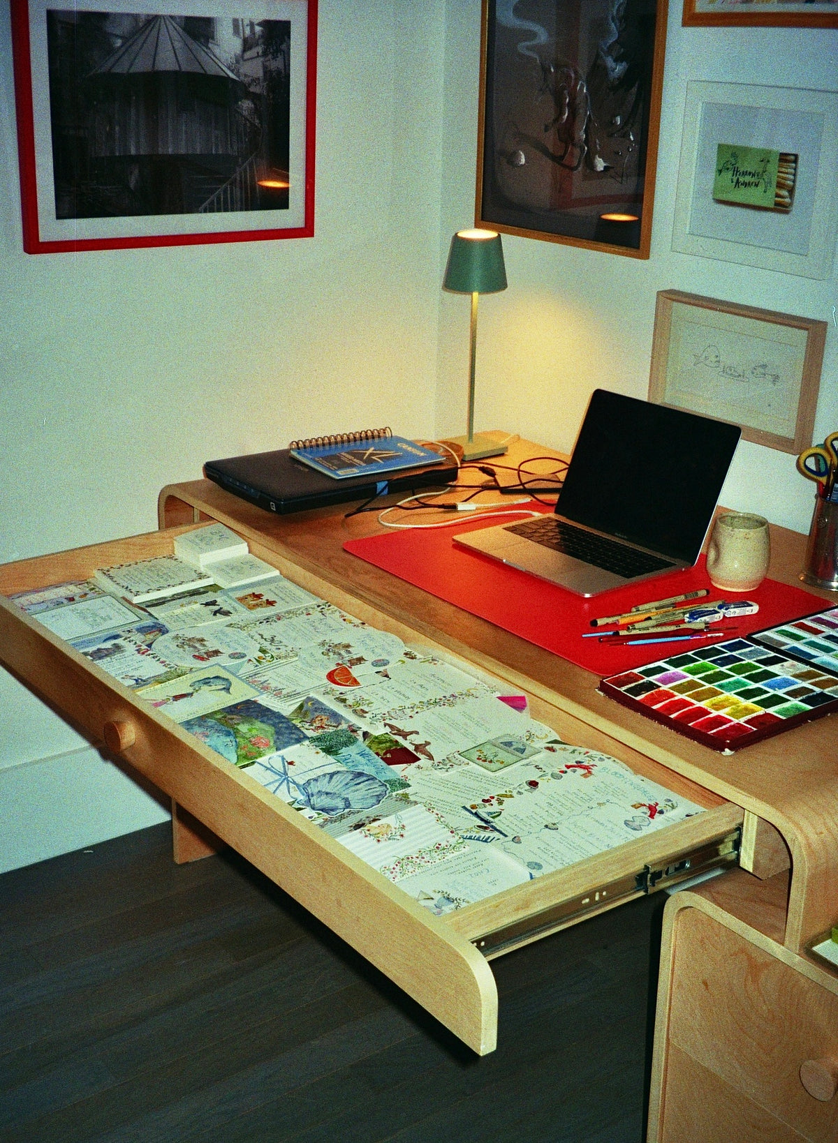 A wooden desk with a pull-out drawer covered in printed pages, shown in a home office setting with a laptop, lamp, framed wall art, and art supplies.