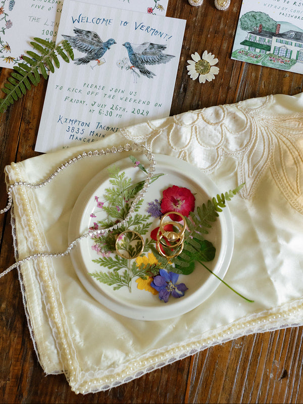 A styled flat-lay of a wedding invitation suite and matching jewelry set on a dark wood background. The set includes a floral invitation card, a welcome card with bird illustrations, a guest book insert, a small illustrated card, a cream lace-trimmed fabric piece, a white plate with edible flowers and rings, and pearl-like bead strands.