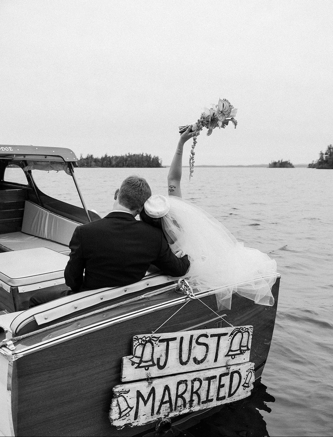 Black and white wedding photograph of a bride and groom sitting in the back of a boat on the water, with the groom holding the bride's hand and a 'JUST MARRIED' wooden sign on the boat's side.
