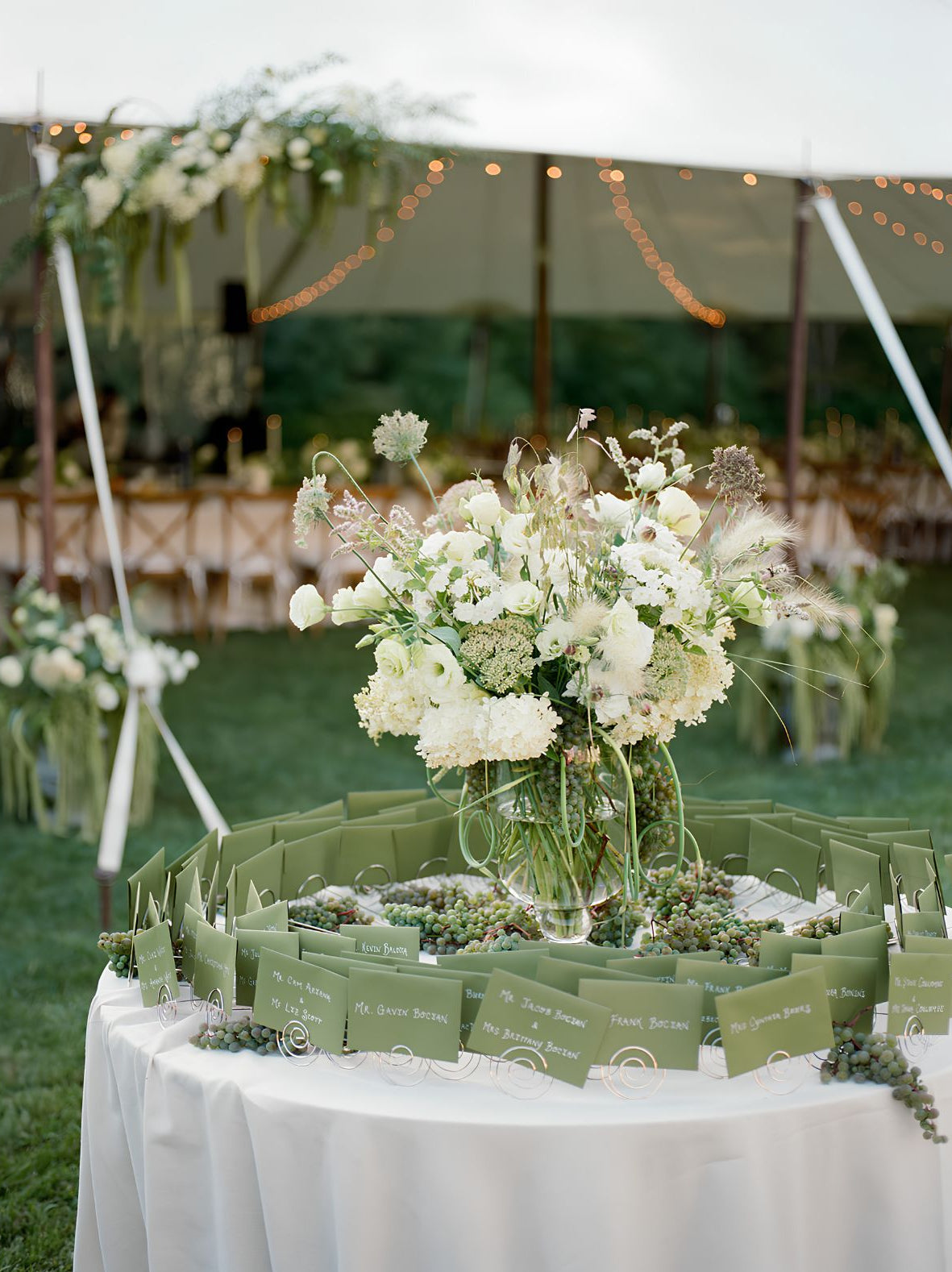 A round table covered in a white tablecloth displays a large floral arrangement in the center and rows of olive green seating cards arranged around the perimeter, set outdoors under a tent with grass and event decor in the background.