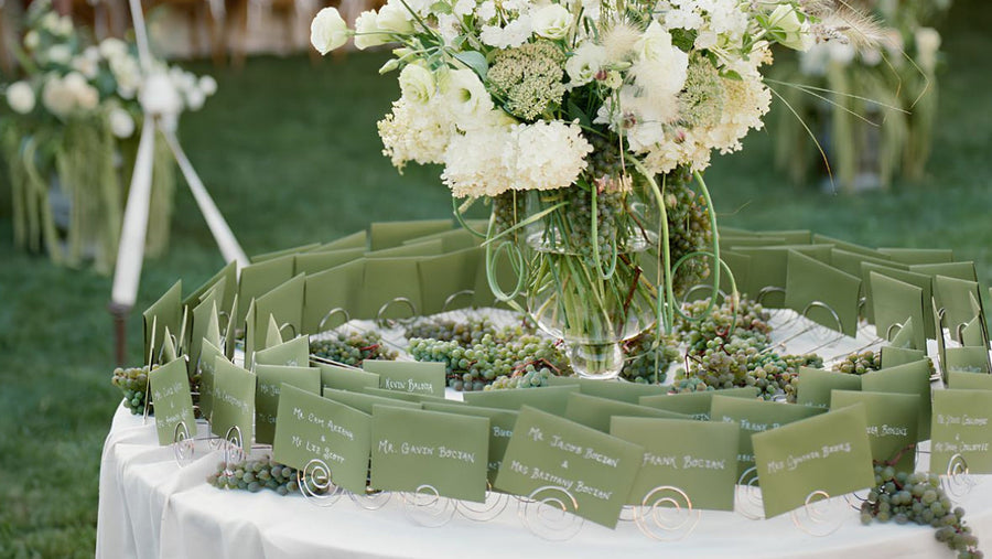 A round table covered in a white tablecloth displays a large floral arrangement in the center and rows of olive green seating cards arranged around the perimeter, set outdoors under a tent with grass and event decor in the background.